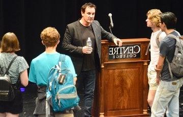 Students carrying backpacks stand in a loose circle around a man holding a water bottle and resting his arm on a podium bearing the Centre College logo. 